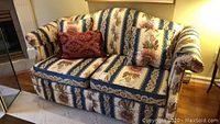 Front view of heritage print fabric loveseat with floral striped pattern and decorative pillows on hardwood floor.