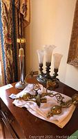 Fluted bulb lamp, silver and gold candle stick, two antique brass candle sconces arranged on a wooden table near curtain and mirror.
