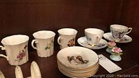 Six bone china cups with floral designs, two cups have blue and pink flowers, others with varied floral patterns, plates with bird design stacked in front.