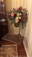 Photo showing metal plant stand with dark bowl vase containing assorted faux flowers and one metal side table with grid pattern glass top.