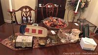 Photo of various items on a wooden table including brass candlestick holders with candles, potpourri bowl, jewelry box, salt and pepper shakers, candy dish, ceramic serving plate, decorative trays, and heated ceramic candle holder