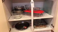 Cupboard shelf with cookware including metal roasting pan, paella pan, glass mixing bowls, Pyrex baking dish, marinade container with lid, cooling rack.