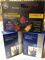 Full box of GE Pro-Line 150 outdoor commercial grade lighting with image showing branding, bulb quantity, and features, along with two Celebrations 100 LED mini light strings boxes stacked in front.