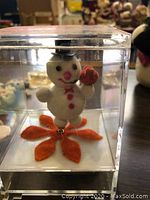 Photo of a vintage snowman music box enclosed in a clear plastic cube, showing the snowman figure with red buttons, a black top hat, and red scarf, standing on an orange leaf-shaped base.
