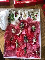 Box containing 16 assorted glass Christmas ornaments wrapped in tissue paper. Includes Santa Claus, snowmen, a Christmas tree, and other themed glass figurines, all hand-painted with colorful and glitter detailing.