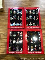 Four red boxes of assorted glass Christmas tree ornaments on wooden table, each containing 12 ornaments with holiday-themed designs including snowmen, nutcrackers, Santa Claus, and more.