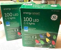 Two unopened green boxes of GE Energy Smart 100 LED C-5 lights for indoor/outdoor use with multicolored bulbs pictured on the packaging.