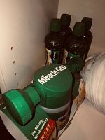 Several bottles of Miracle-Gro liquid fertilizer with green caps and dispensing mechanisms grouped in corner against wall.