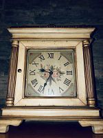 Front view of wooden mantle clock showing the floral face with Roman numeral hour markers and black metal hands, framed by decorative carved columns and distressed cream paint.
