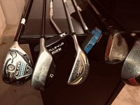Multiple assorted golf clubs resting against a surface, showing a TaylorMade SLDR 5 wood, metallic irons including Bellevue Rescue Mid, and other vintage and metal headed clubs.