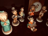 Photo showing several Hummel figurines in various poses on a wooden surface, including a praying child and a boy with a top hat.