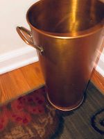 View of full copper umbrella stand showing its height and cylindrical shape placed on wood floor with a rug beneath.