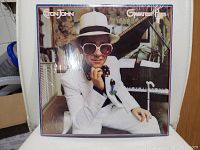 Front cover of Elton John Greatest Hits vinyl record showing Elton John in white suit, sunglasses, and hat seated by piano.