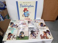Front view of large and 8 smaller Paddington books by Michael Bond laid out on floor showing cover art and titles.