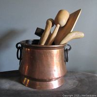 Full front view of the copper pot containing wooden utensils showing overall shape, patina and the rolled top edge.