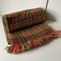 Rolled and folded view of bed coverlet showing fringed edge and plaid pattern with orange, green and black colors.