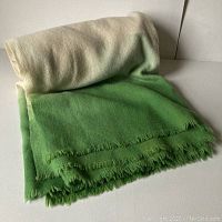 Rolled and folded wool blanket showing cream to green dip dye with fringed edges.
