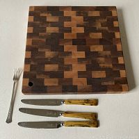 Top and front view of wood butcher block cutting board with 3 knives with yellow Bakelite handles and stainless steel blades plus a silver-plated pickle fork.