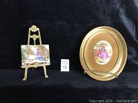 Two vintage porcelain plaques, one large oval with gold border and one small rectangular on brass stand, displayed on black velvet background.