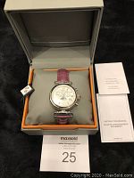 The watch displayed in its orange and grey box, secured around a grey cushion, with tags attached and instruction manuals beside it.