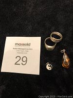 Four pieces of vintage silver jewelry on a black cloth background with auction number card 29: two rings, one silver yin-yang charm, one teardrop pendant with amber-colored center