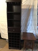 Dark wood wine stand with multiple cubbyholes, shelves, drawer and hooks at top, shown against a backdrop.