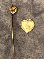 Gold stick pin and 14K gold heart pendant with letter 'A' on a grey cloth background.