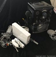 Black karaoke machine with microphone wired, placed beside a white Nintendo Wii console and controllers on a black surface.