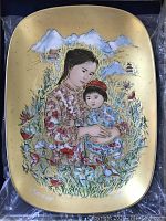 Front view of rectangular yellow plate revealing detailed illustration of mother and child with mountain background and flora. Artist name visible.