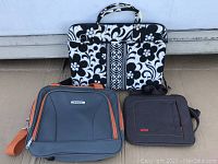 Three electronics carrying bags placed side by side including a patterned fabric handbag, a black and orange backpack style bag, and a small black stm branded bag.