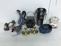 Overview of assorted eclectic decor items on white surface including two duck figurines, tin man wine bottle holder, ceramic pitcher, lantern, candle holder, bowl, and sundial compass.