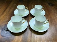 Four white fine bone china espresso cups with gold trim on rims and handles, each paired with matching saucers, arranged on wood surface.