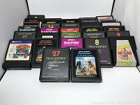 Photo shows multiple vintage Atari 2600 cartridges arranged on a white surface. Visible titles include Superman, Popeye, Raft Rider, Q*bert, Golf, and more.