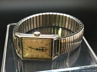 View of the full watch face, showing rectangular beige dial with black Arabic numerals, small seconds subdial, and silver-tone metal case and expandable band.