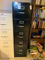 Full view of the black 5 drawer metal filing cabinet next to a beige filing cabinet and bookshelf.