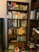 Full front view of tall wood bookcase filled with books and binders, showing shelf layout and overall condition.