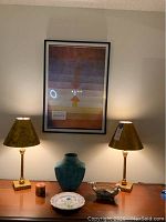 Photo showing the pair of matching lamps with patterned shades turned on, turquoise ceramic vase, framed print by Paul Klee titled 'Separation in the Evening' hung on the wall behind, white decorative ceramic plate with botanical design, and a small candle on wood surface.