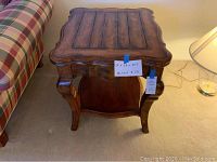 Front view of wood end table showing carved scalloped edges, curved legs, and lower shelf. Blue tape label visible on side.