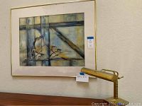 Front view of abstract painting with gold tone frame and adjustable brass lamp beside it.