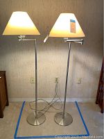 Pair of floor lamps side by side, both with conical lampshades and brushed chrome finish. One lampshade shows visible damage.