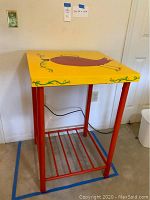 Bar height table showing yellow top with red bird and green vine decoration, red metal legs and lower shelf.