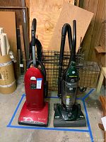 Red Riccar upright vacuum cleaner next to dark green Dirt Devil upright vacuum cleaner with canister and hose attachments, placed on a blue tape-marked floor area in a garage.
