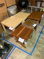 Photo shows five assorted end tables of various wood finishes including two maple laminate and three stained wood with slatted and solid tops. Tables are in a garage setting with tags on each item.