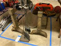 Photo showing two iron andirons, a set of fireplace tools on a stand (poker, brush, shovel), and the metal wood holder in the background.