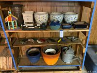 Overall shelving unit showing ceramic pots, bird feeder, handspreaders, watering can, garden tools, and sprinklers arranged on shelves