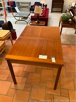 Photo of the teak dining table showing the top surface with closed leaf extension, in a room setting with other furniture visible.