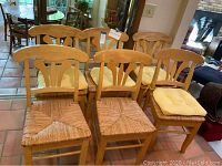 Wide view of all six wooden dining chairs, showing woven seats and yellow seat pads on four chairs.