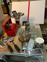 Full overview of kitchenware items on table including cheese baker, lemon juicer, utensils, butter dish, measuring cups and small accessories.