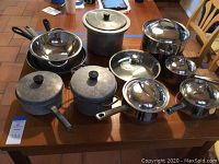 Full view of all assorted cookware including stainless steel and vintage aluminum pots and pans with lids on a wooden table.