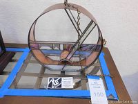 View of the front of the circular stained glass and metal sculpture showing geometric colored glass pieces inside metal frame and attached chain.